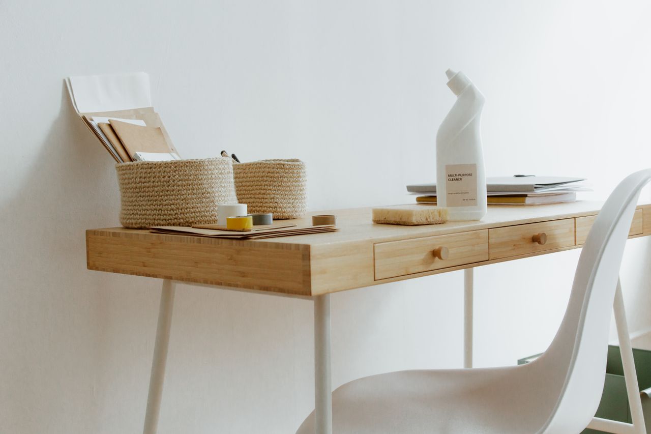 gallery-02 A clean and organized home office desk with cleaning products, papers, and storage baskets.