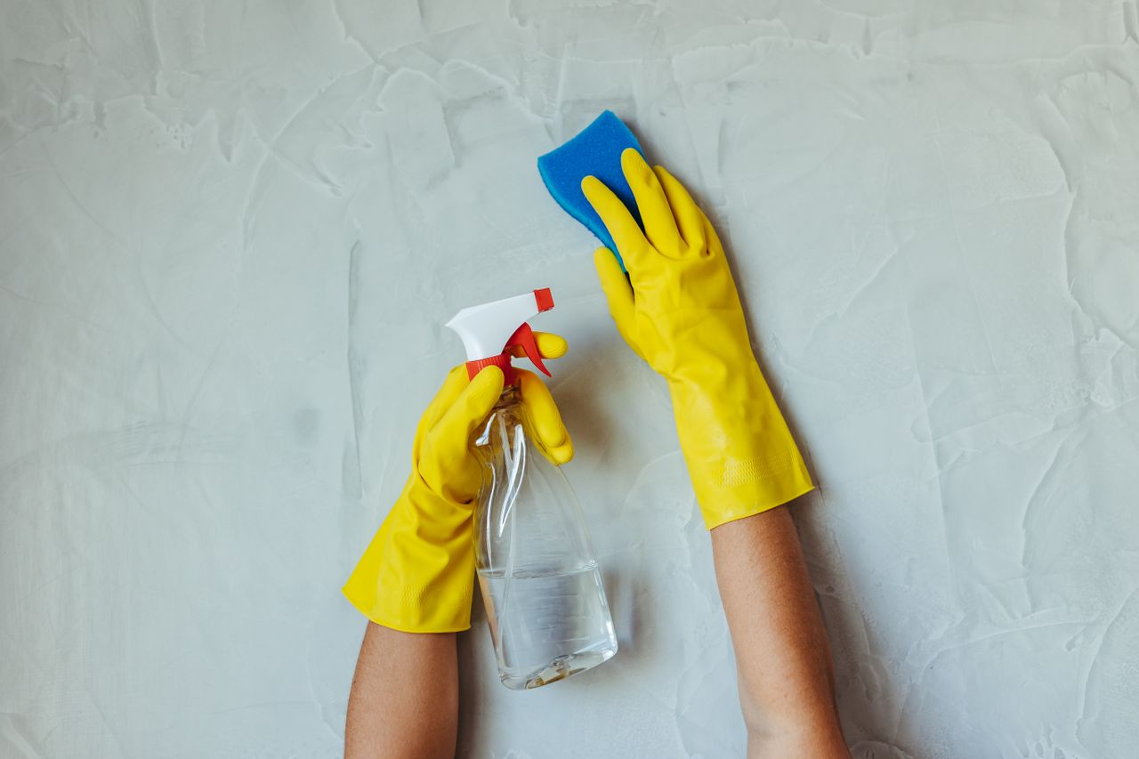 services-04 Gros plan sur des mains gantées de jaune nettoyant un mur à l'aide d'un flacon pulvérisateur et d'une éponge.